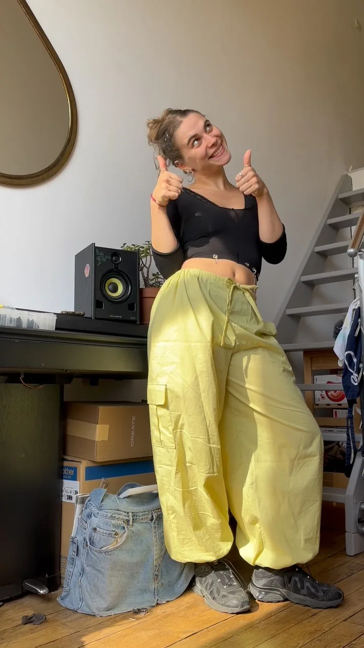 Lola, pouces levés, en pantalon parachute jaune dans son atelier